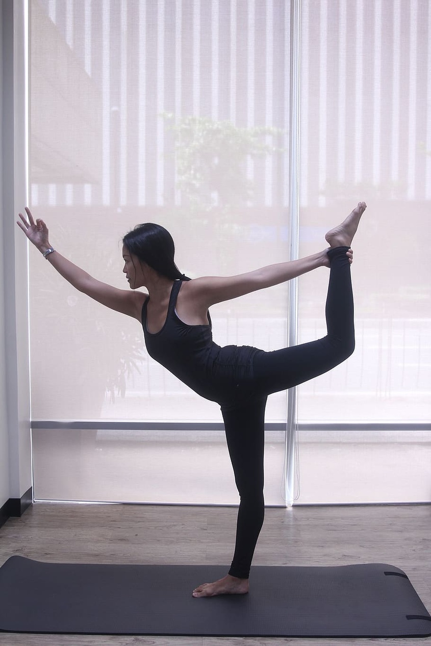 En kvinna i svarta träningskläder utför dansarens position (Natarajasana) på en yogamatta i en ljus studio, vilket visar en kombination av yogateknik och styrka.